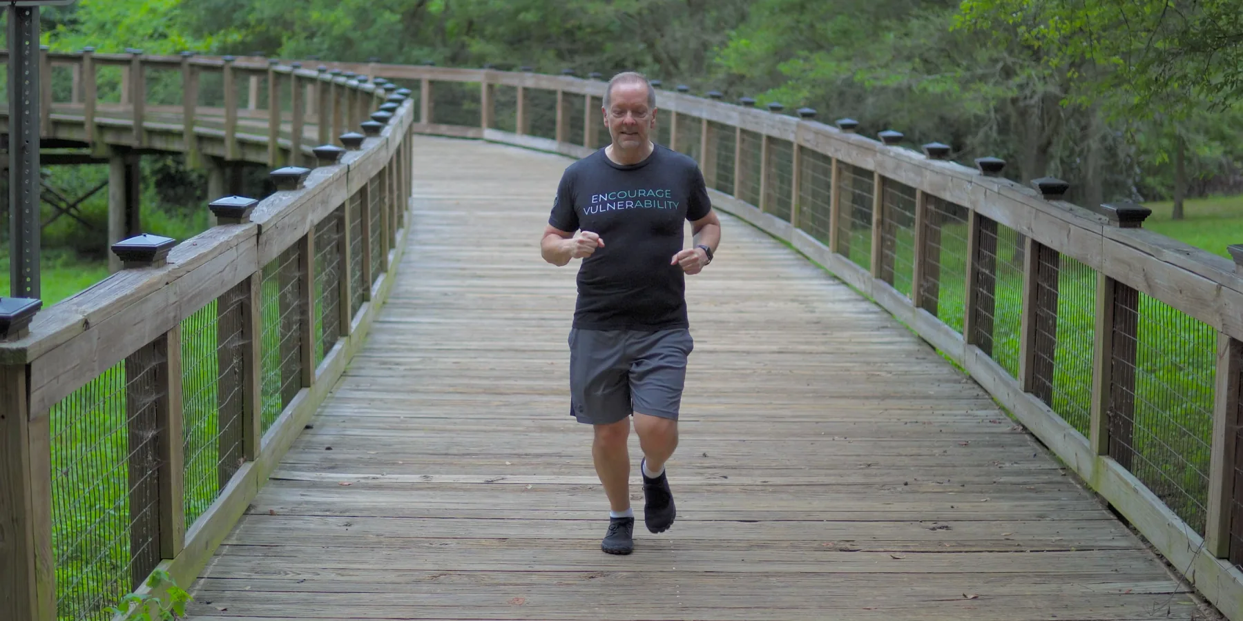 A man running at a steady pace along a flat, measured boardwalk through green woods