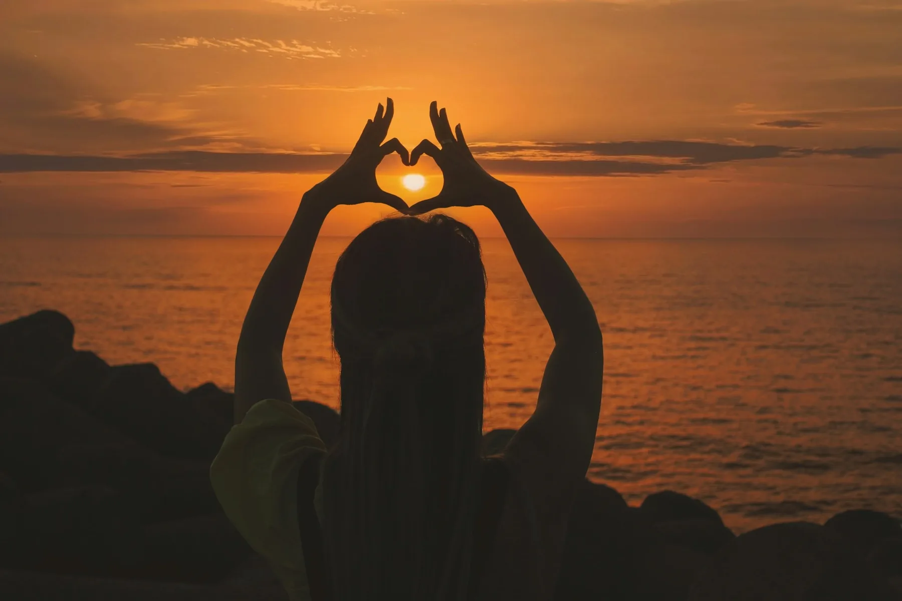 A person at the beach at sunset framing the setting sun inside a heart made with their hands