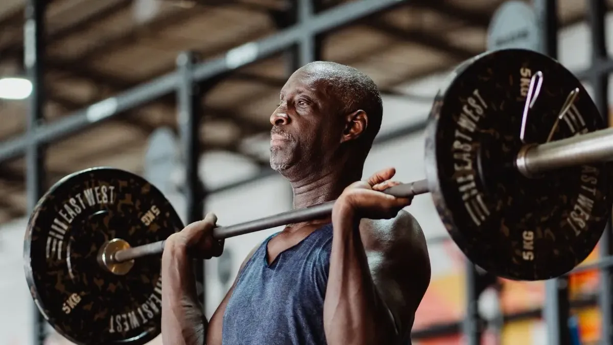 An older man holding a loaded barbell at his shoulders in a gym