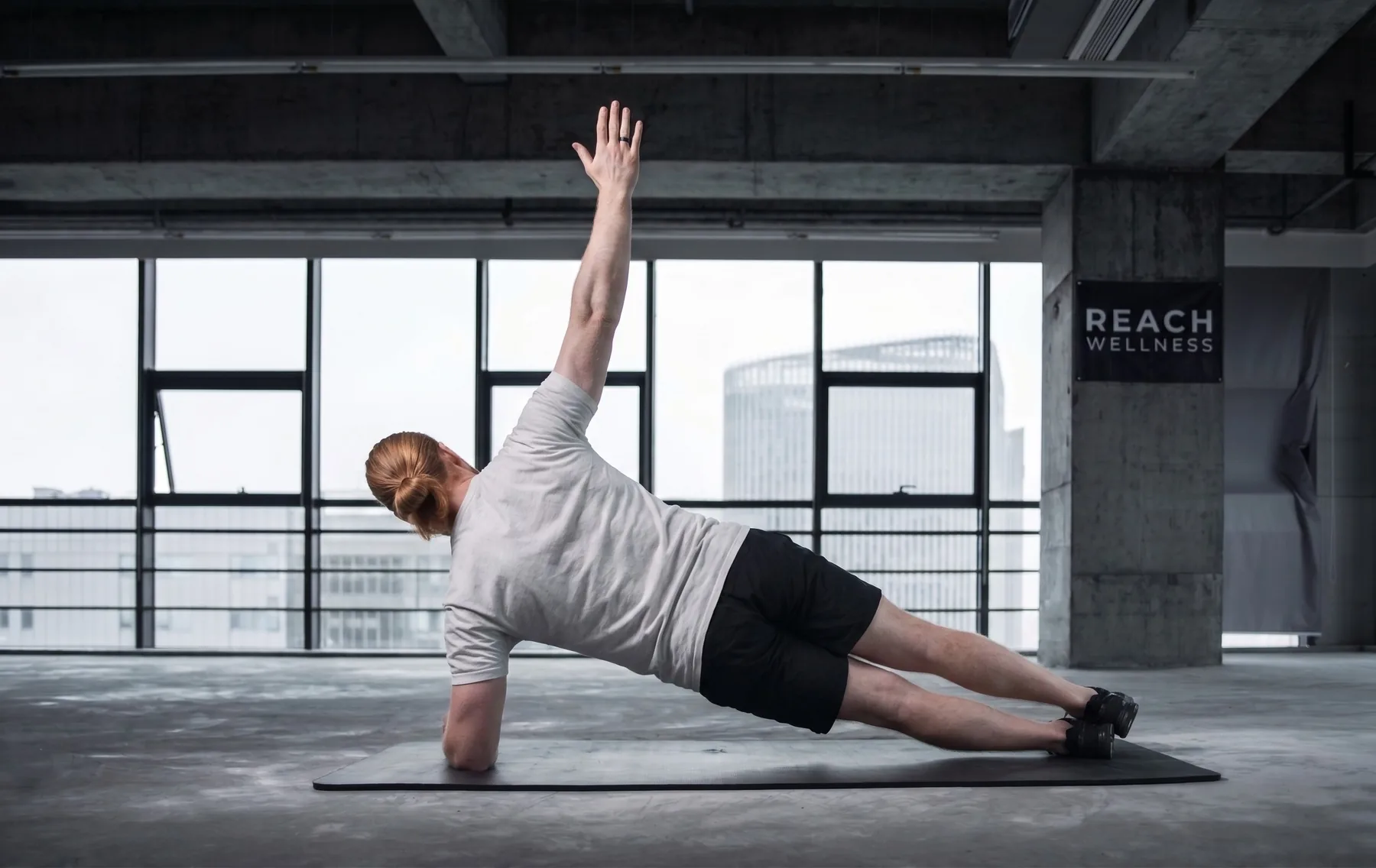 A man holding a side plank on a mat in a bright, open studio, his top arm reaching straight toward the ceiling